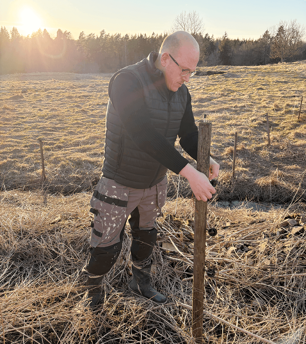 En person med glasögon, svart väst och kamouflagebyxor står på ett fält i solnedgången och trycker ner en träpåle i marken med båda händerna. Torrt gräs täcker marken och träd syns i bakgrunden.