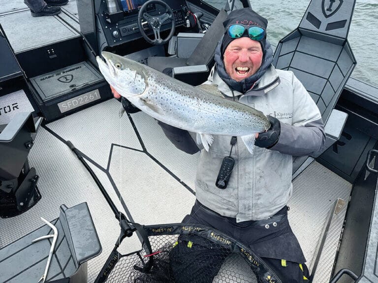 En person står på knä på en båt och håller en stor fisk med båda händerna. Personen har på sig en grå jacka, svarta byxor, handskar och en hatt med solglasögon på toppen. Båtutrustning och vatten syns i bakgrunden.