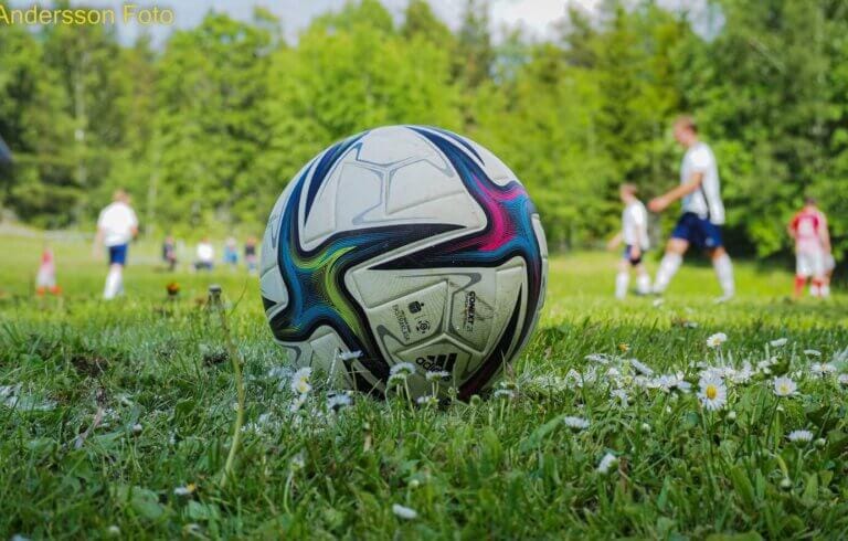 Närbild av en fotboll som vilar på gräs med små blommor. I bakgrunden spelar flera personer i sportuniformer fotboll på en plan omgiven av träd, vilket fångar den livliga andan i fotbollssäsongen.