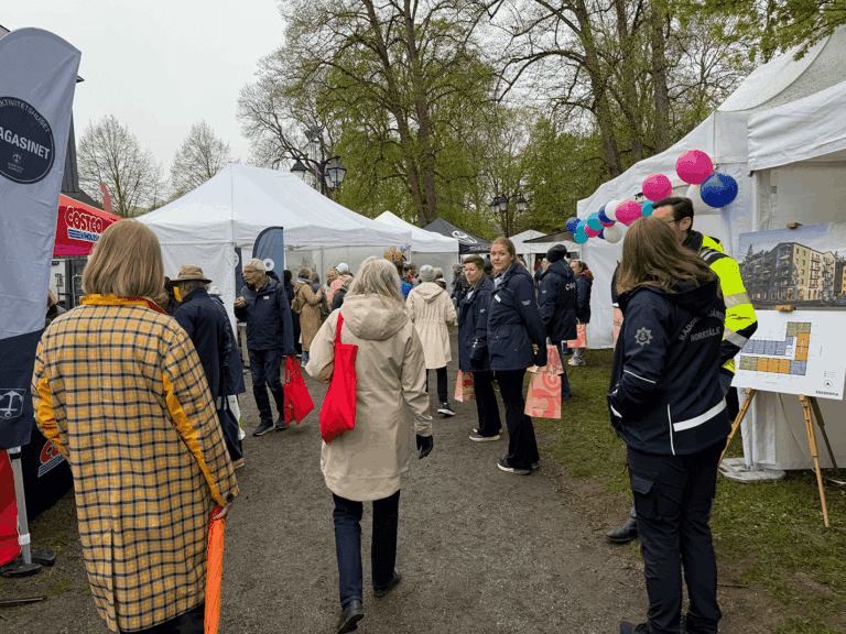 Människor går längs en stig som kantas av utomhustält vid ett evenemang. Några bär jackor och väskor. Ballonger och en tavla med en byggnadsplan är synliga. Scenen utspelar sig i en park en molnig dag.