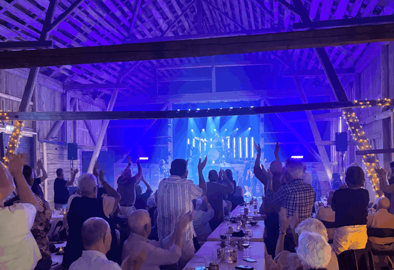 Publiken i en rustik lokal i ladugårdsstil tittar på ett liveband som uppträder på en scen med blå och lila belysning; många står upp och klappar händerna, och i förgrunden står bord dukade för middag.