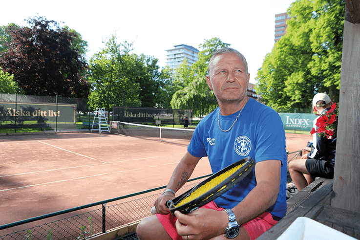 En man i blå skjorta och röda shorts sitter på en bänk med ett tennisracket i handen bredvid en grusbana, med träd och byggnader i bakgrunden. Vädret ser soligt och klart ut.