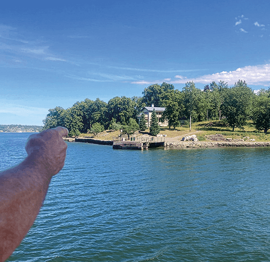 En persons arm sträcker sig ut från bildens vänstra sida och pekar mot ett hus omgivet av träd vid en gräsbevuxen strand, med lugnt blått vatten i förgrunden och en klar himmel ovanför.