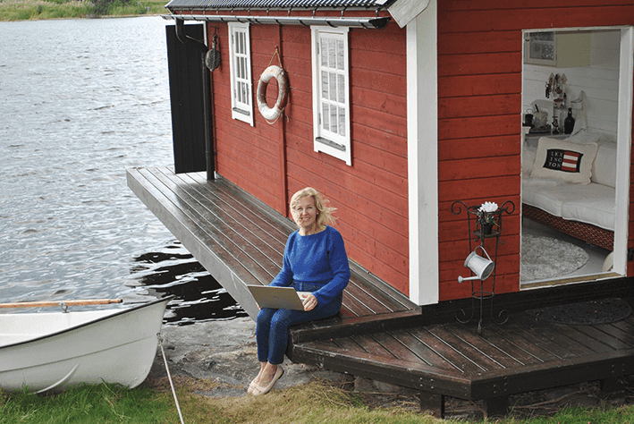 En kvinna sitter på trätrappan till en röd stuga vid sjön med en bärbar dator i knät. En vit roddbåt ligger förtöjd i närheten, och stugans dörr står öppen och avslöjar en mysig interiör. Det verkar blåsa en lätt bris.