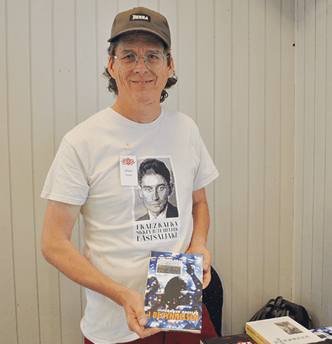 En person med glasögon, keps och en T-shirt med tryck står inomhus, ler och håller i en bok med ett färgglatt omslag. En namnskylt syns på tröjan, och framför personen står ett bord med fler böcker.