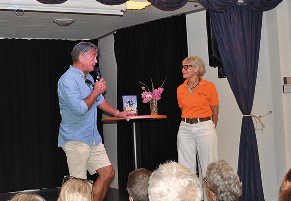En man med en mikrofon talar till en kvinna i en orange tröja och vita byxor på en liten scen. På ett högt, runt bord mellan dem står en vas med rosa blommor och en bok. I förgrunden syns personer ur publiken.