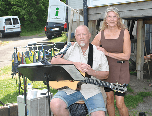 En man sitter och håller i en akustisk gitarr, med ett notställ framför sig. En kvinna står bredvid honom med handen på hans axel. De befinner sig utomhus nära en byggnad och flera flytvästar hänger i närheten.
