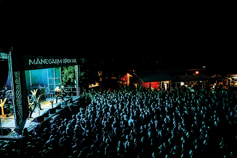 En stor folkmassa tittar på ett liveframträdande på en utomhusfestival som hålls på kvällen. Scenen är upplyst av grönt ljus och har en banderoll med texten ”Månegarm Open Air”. Publiken sitter vänd mot scenen, och i bakgrunden syns några tält.