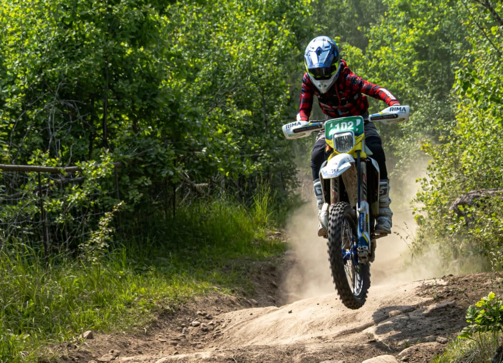 En motocrossförare i en röd rutig skjorta och blå hjälm kör en terrängmotorcykel med startnummer 102 längs en grusväg med grönska och träd på båda sidor. Damm virvlar upp bakom motorcykelns bakhjul när Wiggo Lifvendahl – känd för sina SM-segrar – med säker hand tar sig fram längs banan.