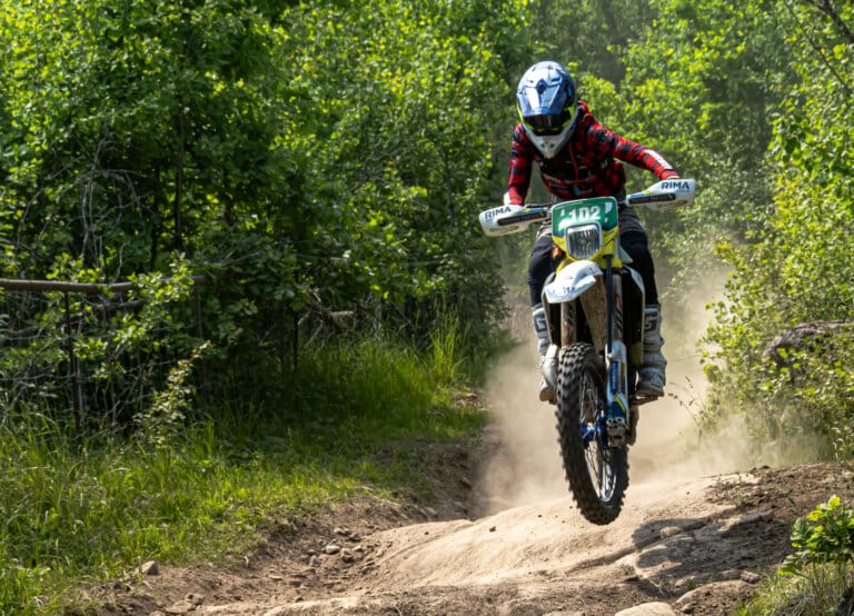 En motocrossförare i en röd rutig skjorta och blå hjälm kör en terrängmotorcykel med startnummer 102 längs en grusväg med grönska och träd på båda sidor. Damm virvlar upp bakom motorcykelns bakhjul när Wiggo Lifvendahl – känd för sina SM-segrar – med säker hand tar sig fram längs banan.