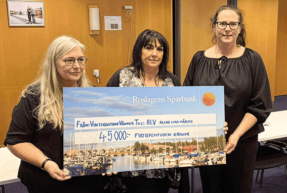 Tre kvinnor står inomhus och håller upp en stor symbolisk check på 45 000 kronor från Roslagens Sparbank. I bakgrunden syns träpaneler och en skärm. På checken avbildas båtar och en scen vid vattnet.