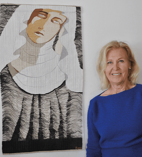 En kvinna med axellångt blont hår, klädd i en blå tröja, står bredvid ett textilkonstverk som föreställer en stiliserad figur med ett neutralt ansiktsuttryck och draperade kläder i beige, svarta och vita nyanser.