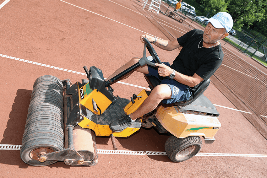 En man i keps och shorts kör en gul vält på en lertennisbana och packar ytan. I bakgrunden syns vita linjer och tennisnät.