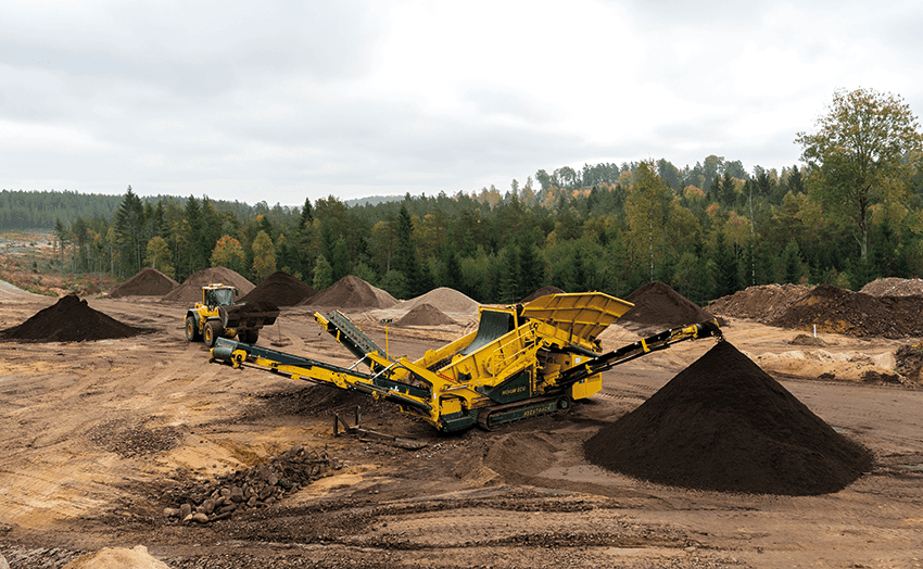 En gul industrimaskin och en lastmaskin arbetar på en jordplan, där de sorterar och staplar jord i högar. I bakgrunden syns skogsklädda kullar och en molnig himmel. Scenen verkar utgöra en del av en gruvdrift eller en markarbetsoperation.
