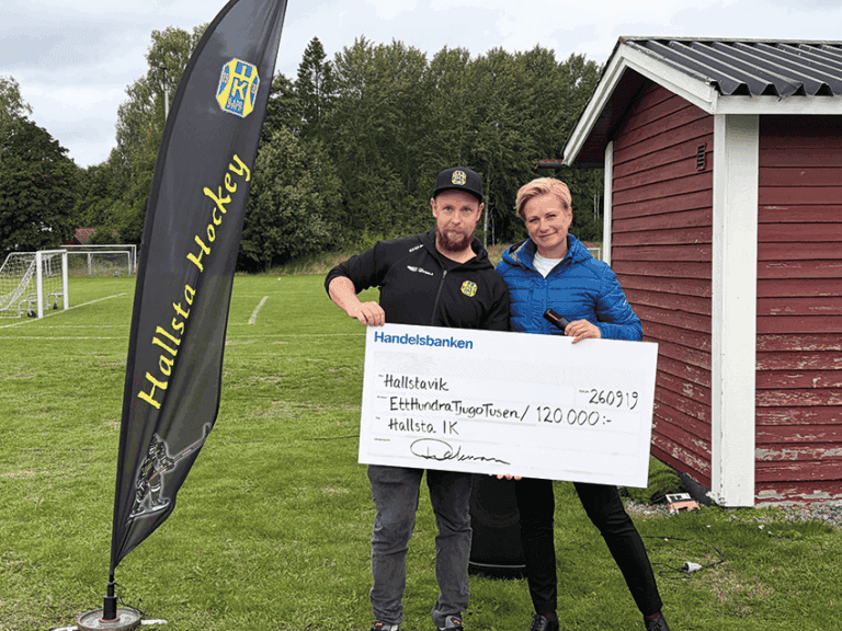 Två personer står utomhus på gräset och håller i en stor ceremoniell check på 120 000 SEK utställd till Hallsävik. En Hallsta Hockey-banner och en röd byggnad syns i bilden. I bakgrunden syns fotbollsmål och träd.