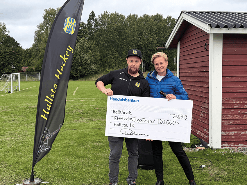 Två personer står utomhus på gräset och håller i en stor ceremoniell check på 120 000 SEK utställd till Hallsävik. En Hallsta Hockey-banner och en röd byggnad syns i bilden. I bakgrunden syns fotbollsmål och träd.