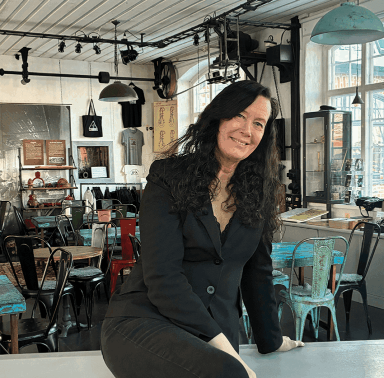 En kvinna med långt mörkt hår, klädd i en svart kavaj och byxor, sitter på ett bord på ett kafé med en eklektisk inredning, där man i bakgrunden kan se stolar som inte matchar varandra och industriell belysning.