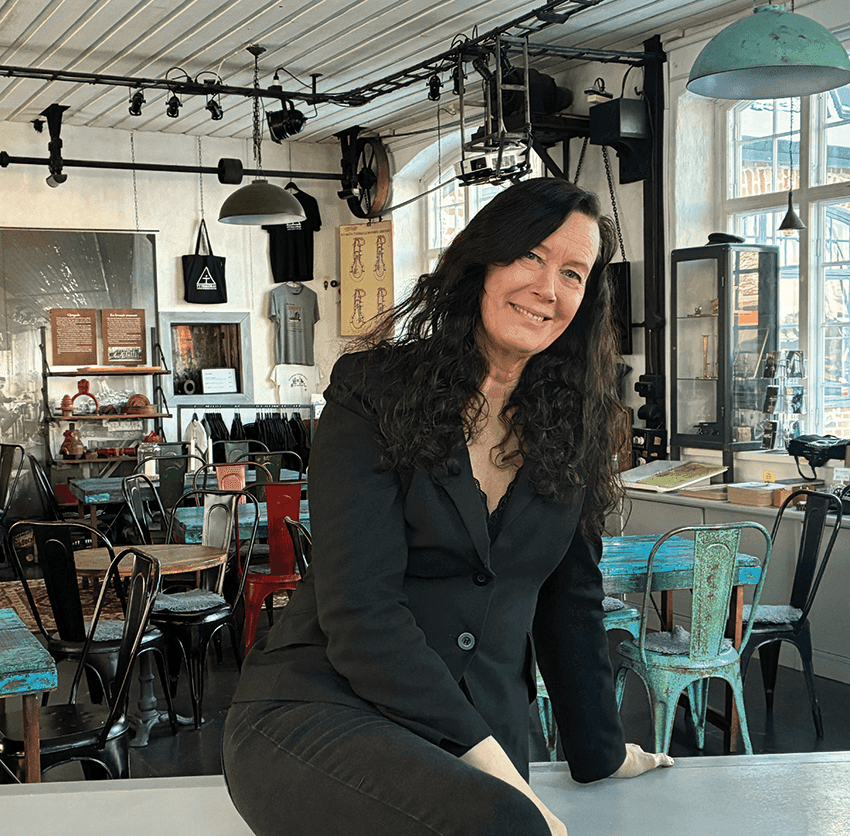 En kvinna med långt mörkt hår, klädd i en svart kavaj och byxor, sitter på ett bord på ett kafé med en eklektisk inredning, där man i bakgrunden kan se stolar som inte matchar varandra och industriell belysning.