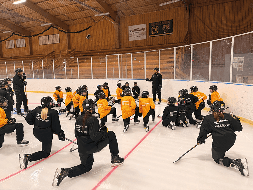 En grupp unga ishockeyspelare i orange och svarta tröjor knäböjer på isen runt en tränare i en inomhushall med träbänkar och väggar. Tränaren verkar ge instruktioner.