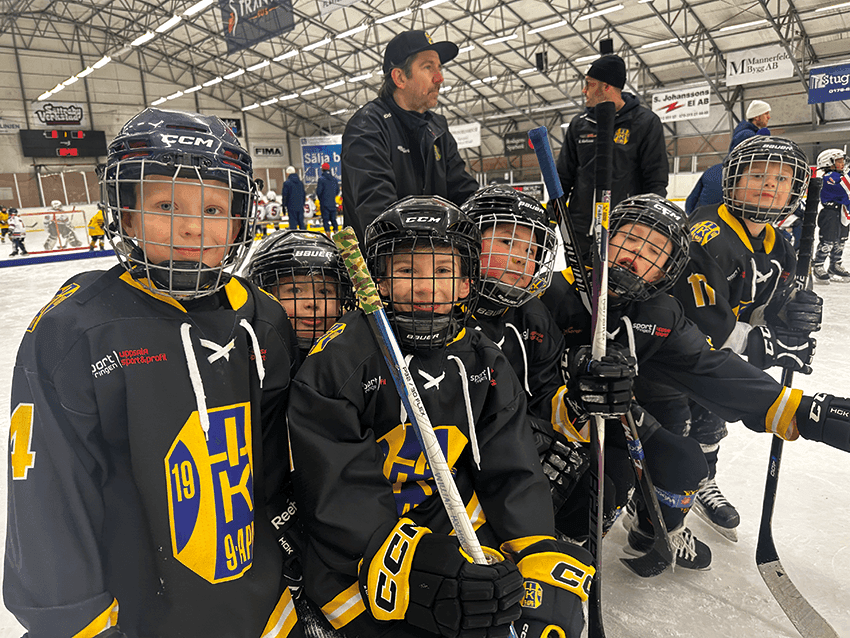 Sex unga ishockeyspelare i svarta och gula tröjor poserar tillsammans på isen med sina klubbor i händerna. En vuxen tränare står bakom dem. Scenen utspelar sig i en inomhushall där man kan se andra människor i bakgrunden.