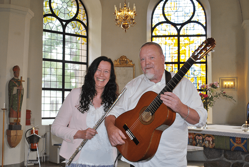 En kvinna med en flöjt och en man med en gitarr står tillsammans i en kyrka. De ler båda två. I bakgrunden syns glasmålningar och en ljuskrona.