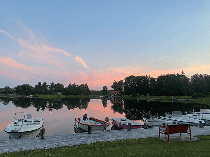 Flera små båtar ligger förtöjda längs en träbrygga vid en lugn sjö i solnedgången. Himlen och träden speglas i vattnet. I förgrunden syns gräs och en röd grill.