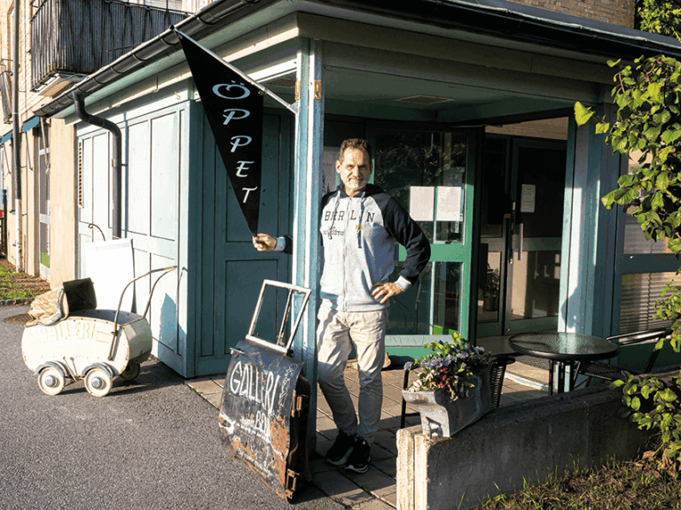 En man står utanför ingången till en byggnad och håller i en skylt med texten ”ÖPPET”. I närheten står en liten vagn med texten ”GALLERI” och några krukväxter. Solen kastar skuggor över scenen.