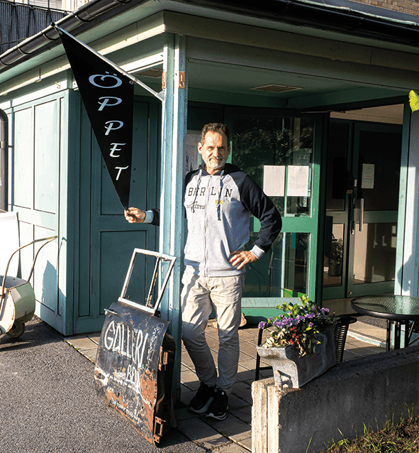 En man står utanför ingången till en byggnad och håller i en skylt med texten ”ÖPPET”. I närheten står en liten vagn med texten ”GALLERI” och några krukväxter. Solen kastar skuggor över scenen.