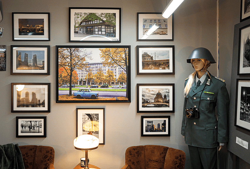 En vägg med inramade foton, en gammaldags lampa, två bruna stolar och en skyltdocka i en grön militäruniform med hjälm och en kamera hängande runt halsen. En stark taklampa syns delvis uppe till höger.