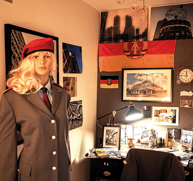 En skyltdocka klädd i en grå uniform och en röd basker står i ett hörn bland tyska flaggor, inramade foton och ett rörigt skrivbord med en lampa som sprider ett varmt ljus. Scenen påminner om ett litet, inrett kontor eller arbetsrum.