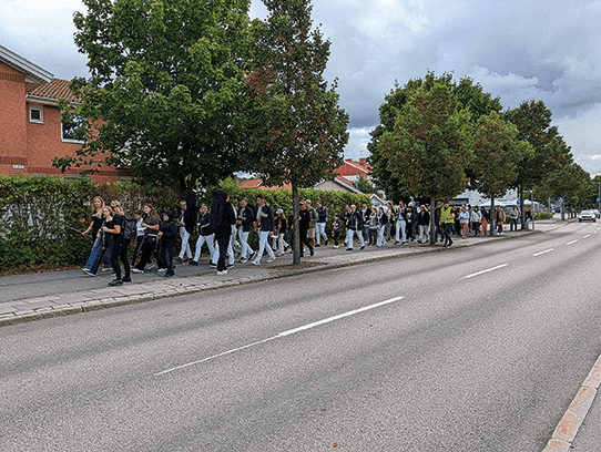 En grupp människor går längs en trottoar intill en väg kantad av träd och byggnader. Himlen är molnig och gatan verkar tom på fordon.