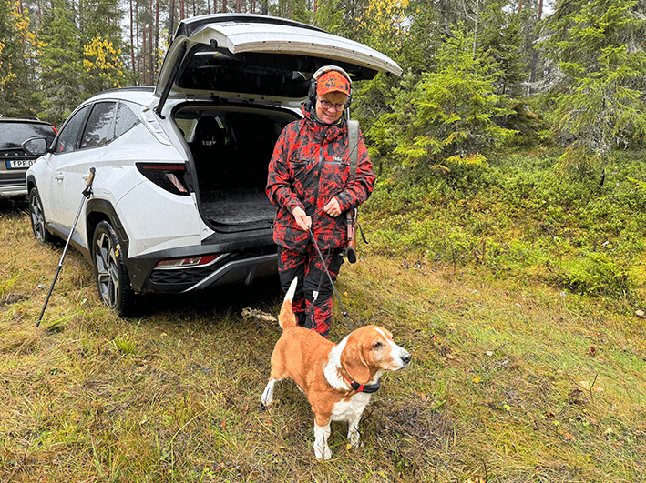 En person klädd i kamouflagejaktkläder står vid den öppna bagageluckan på en vit SUV med en beagle i koppel i en skog.