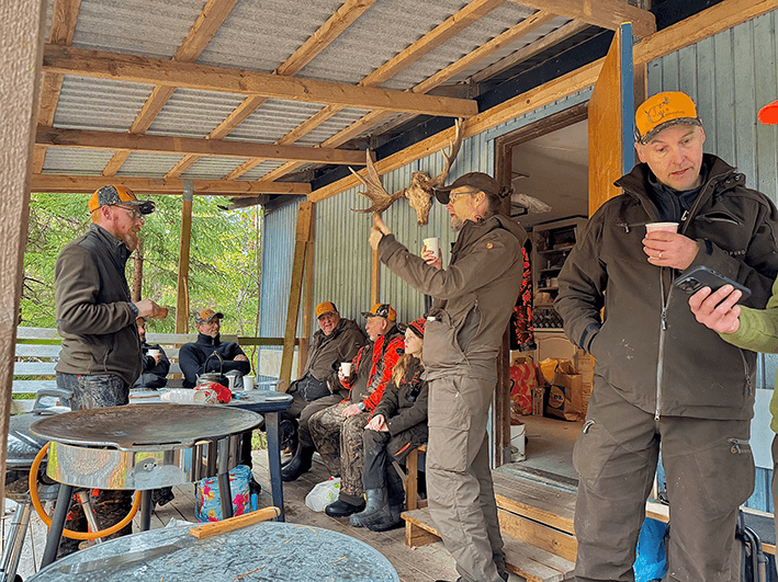 En grupp människor klädda i friluftskläder och kepsar samlas på en träveranda, dricker ur muggar och pratar.