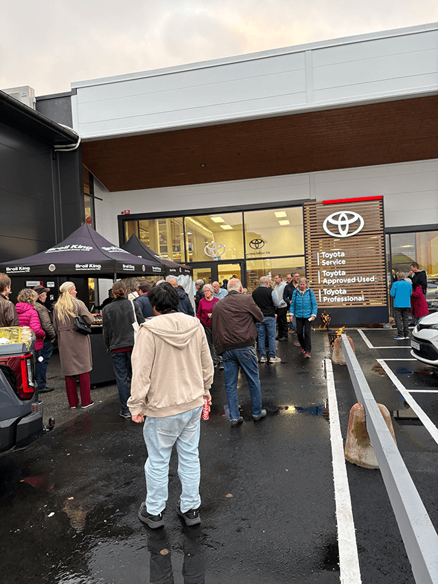 En grupp människor som står utanför serviceingången till en Toyota-återförsäljare bredvid ett tält med Broil King-logotypen.