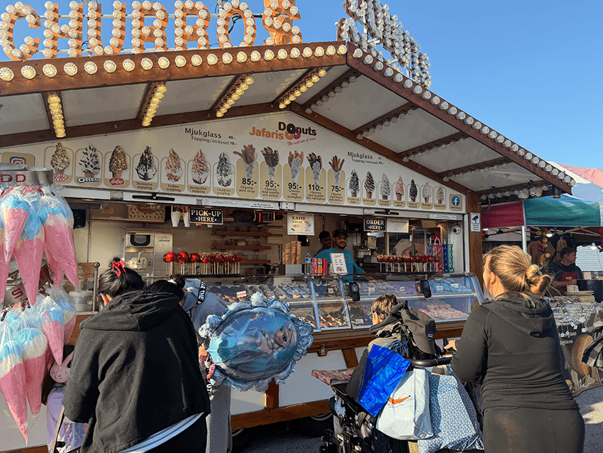 Ett matstånd som säljer churros, glass och munkar. Folk står vid disken, däribland en kvinna med en barnvagn och en annan med ballonger och sockervadd. Menyn och olika sötsaker syns i utställningen.