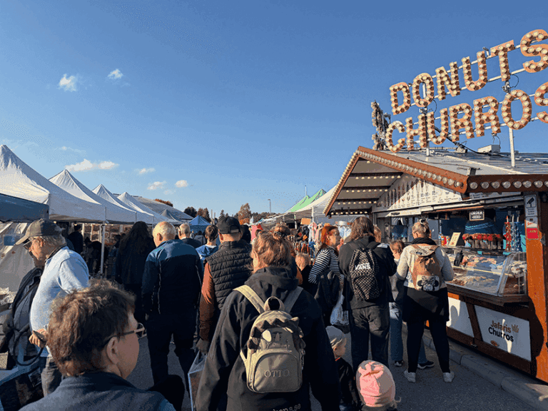 Folk går genom en utomhusmarknad med tält och en matbod som säljer munkar och churros. Himlen är klar och blå. Folkmassan verkar titta runt eller stå i kö vid marknadsbodarna.