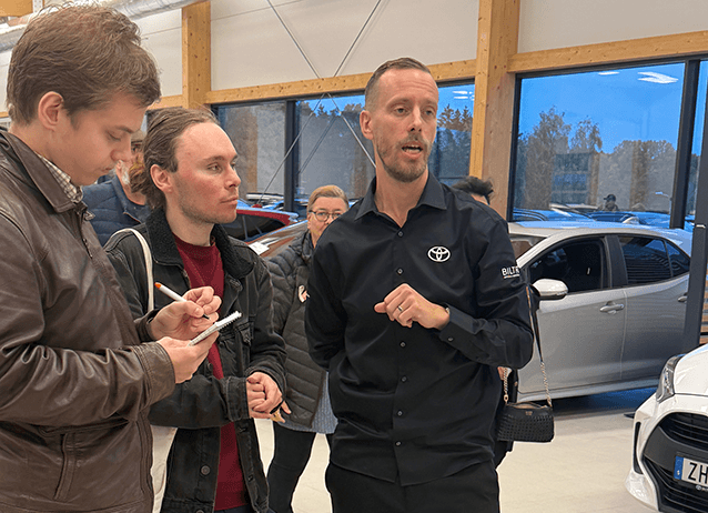 En man i en svart Toyota-tröja pratar med två män som antecknar i en bilhall.