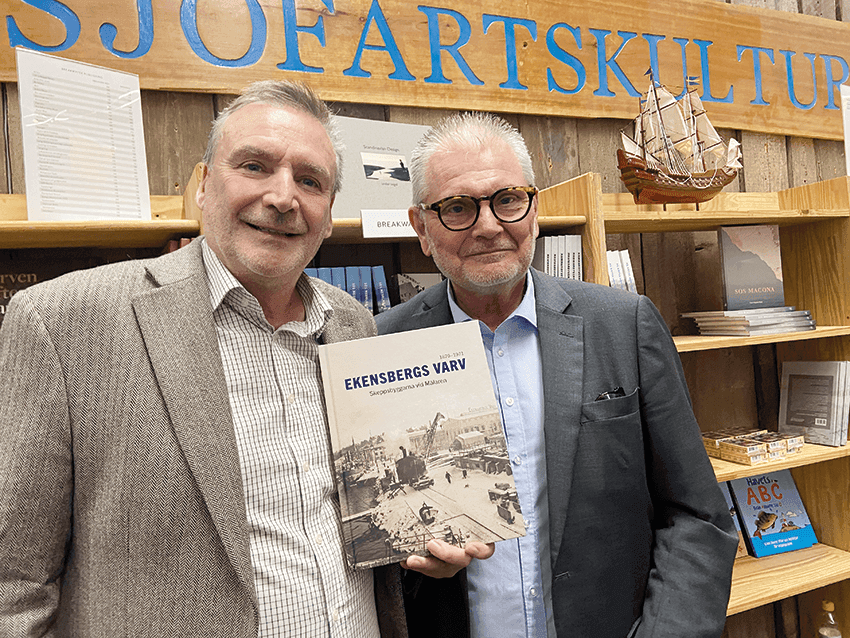 Två män står framför en bokhylla; den ene håller i en bok med titeln ”Ekensbergs Varv”.